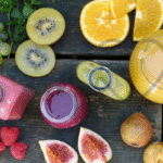 A top view of assorted fresh fruits including kiwi, oranges, figs, raspberries, grapes, and various colorful fruit juices in bottles and a jug on a rustic wooden table.