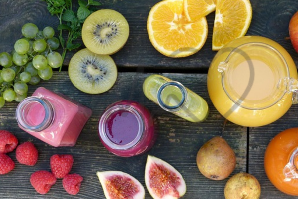 A top view of assorted fresh fruits including kiwi, oranges, figs, raspberries, grapes, and various colorful fruit juices in bottles and a jug on a rustic wooden table.