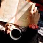 A person reading a book while holding a cup of coffee, wearing a red watch, with warm sunlight falling on the pages.
