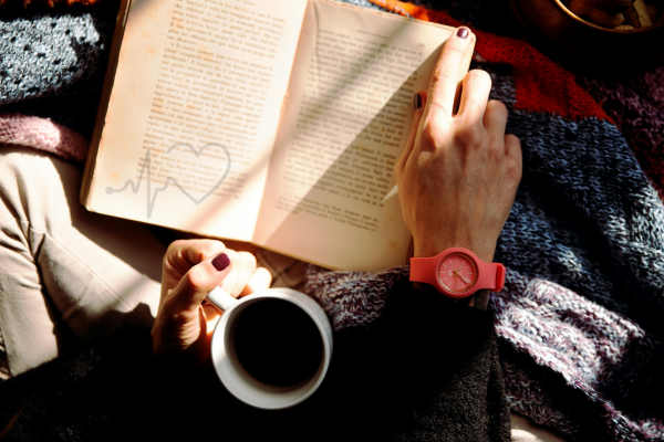 A person reading a book while holding a cup of coffee, wearing a red watch, with warm sunlight falling on the pages.