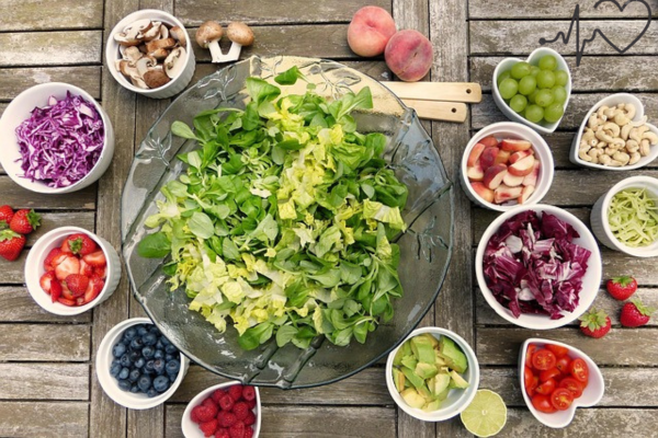 A large glass bowl filled with mixed green salad, surrounded by smaller bowls containing various colorful vegetables and fruits on a wooden table.